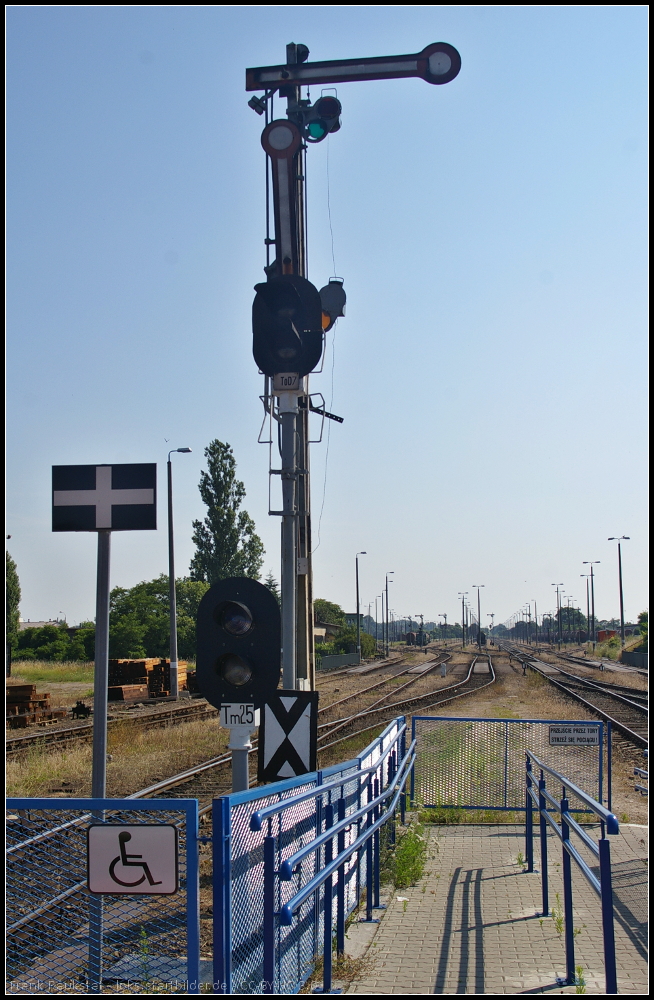 Drei Signale, ein Warnschild und der ungesicherte barrierefreie bergang von Peron 2 zu Peron 1 am Bahnhof Kostrzyn. Das Formsignal ist auer Betrieb und nicht wie in Deutschland blich entsprechend gekennzeichnet. Davor steht das Signal ToD7, davor Tm25 mit blauem Signalbild und dann das typische Hinweisschild fr Fugnger (weies Kreuz auf schwarzem Grund).