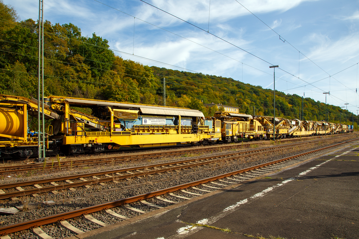 
Bild 1
Die Plasser & Theurer Bettungsreinigungsmaschine RM 95-800 W der MGW Gleis- und Weichenbau-Gesellschaft mbH & Co. KG (Berlin) ist am 20.09.2020 im Bahnhof Dillenburg abgestellt.

Hier ein Überblick über die eigentliche Maschine. Von links nach rechts die Abwasserkläranlage 99 80 9510 002-5 D-MGWB und die eigentlichen Bettungsreinigungsmaschine RM 95-800 W (99 80 9414 001-4 D-MWGB), sowie am Zugende noch der Schutzwagen 3999 010-6 der Gattung Res.

Die Grundmaschine wurde 2010 von Plasser & Theurer unter den Fabriknummern 5221 bis 5224 gebaut, sie hat die VDM Nr. 99 80 9414 001-4 D-MWGB.

Bei der RM 95-800 W von Plasser & Theurer handelt es sich um eine Hochleistungsbettungsreinigungsmaschine mit integriertem Schotterwaschmodul und einer Abwasserkläranlage.

Im Rahmen der kompletten Schotteraufbereitung wird das sogenannte Feinkorn separiert und nur noch dieses muss abtransportiert werden. Der zur Wiederverwendung vorgesehene Schotter wird gewaschen, wieder angeschärft und anschließend in das Gleisbett eingebracht. Durch diese Maßnahmen werden eine bessere Qualität des Schotterbetts und eine längere Liegedauer gewährleistet. Optional kann jedoch auch Neuschotter zugemischt werden. Durch diese besondere Konzeption der Maschine tritt nicht nur eine erhebliche Kosteneinsparung ein, sondern auch die mit dem geringeren Transportaufwand verbundenen Emissionen werden erheblich reduziert. 

TECHNISCHE DATEN der RM 95-800 W: 
Gesamtgewicht: 427.000 kg 
Länge über Puffer: 114.980 mm (ohne Klärwagen) 
Drehzapfenabstände: 15.000/ 25.500/ 14.500/ 16.000/10.000/10.500 mm 
Höhe: 4.295 mm
Breite: 3.250 mm
Achsanzahl: 22
Höchstgeschwindigkeit : 19 km/h (Eigenfahrt) / 100 km/h (geschleppt):
Reinigungsleistung: bis 300 m³/h bei Reinigung/Vollaushub und Waschbetrieb.