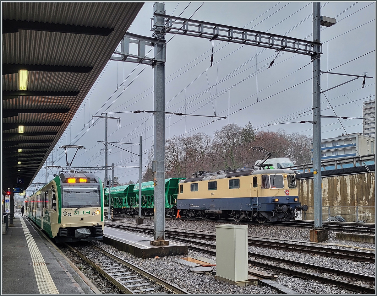 Da die BAM /MBC eigene Re 4/4 II 506 (Re 420 506) zur Zeit in der Revision in Bellinzona weilt, hat die BAM MBC die IRSI/IGE  Rheingold  Re 4/4 II 11387 (Re 421 387-2) angemietet. Die Lok trägt die UIC Nummer Re 421 91 885 4421 387-2 CH-IRSI. Das Bild die Lok nachdem sie ihre Wagen des Güterzugs Gland - Apples auf die Rollbockanlage in Morges geschoben hat und nun Abgekuppelt wird. 
Links im Bild ein BAM Regio R 76 nach Bière.

22. Februar 2024