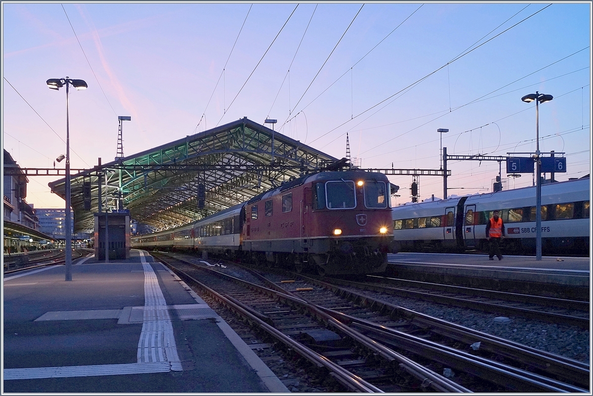 Ein leichtes Morgenrot breitet sich über dem noch im Schatten liegenden Bahnhof von Lauanne aus, im Vordergrund ist eine SBB Re 4/4 II mit ihrem RE aus St-Maurice eingetroffen. 

20. Feb. 2019