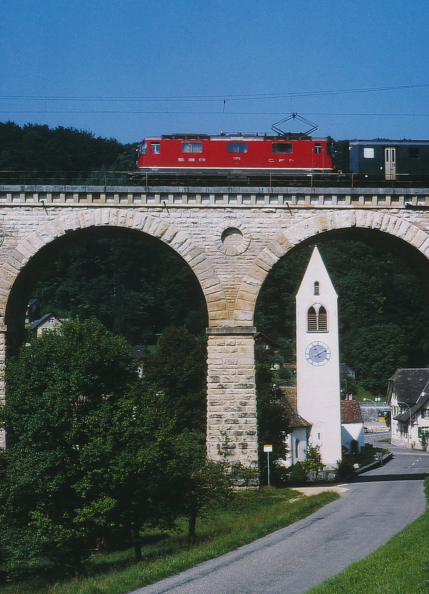 SBB: Obschon in der Schweiz jeden Tag sehr viele Re 4/4 mit Reise- Güter- und Postzügen unterwegs sind, handelt es sich hier um eine sehr seltene Aufnahme. Zur Abwechslung konnte im Sommer 1996 eine Re 4/4 I der zweiten Serie mit einem umgeleiteten Reisezug vor der herrlichen Kulisse der Kirche Läufelfingen beim Passieren des Viadukts verewigt werden.
Foto: Walter Ruetsch  