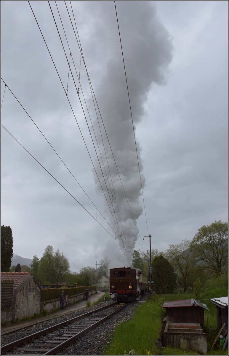 Vapeur Val-de-Travers: Train  Au fil de l'Areuse .

Während mit dem Fahrdienstleiter telefoniert werden musste, hatte E 3/3 8511 einen Hauch zu viel Kohlen aufgelegt bekommen. Mit dem Regenwetter ist es allerdings beeindruckend. Die Weiterfahrt konnte doch noch beginnen und der Fotograf mutierte vorübergehend zum Bahnübergangssicherungsposten. Môtiers, Mai 2023.