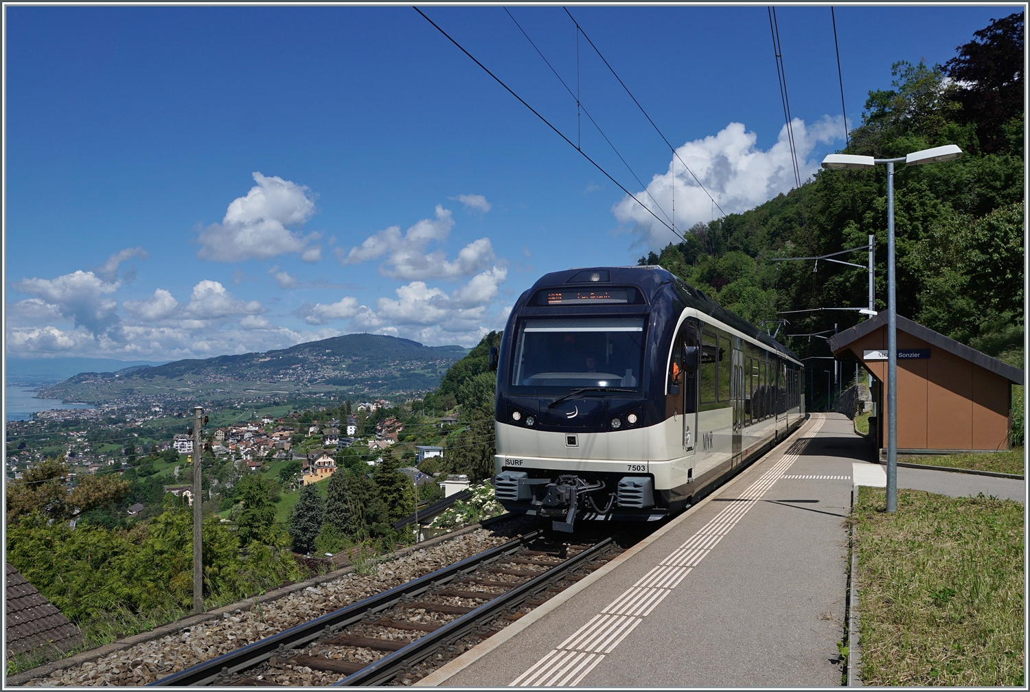 Bild mit Aussicht: Während der CEV MVR ABeh 2/6 7503   Blonay-Chamby  als Regionalzug von Montreux nach Les Avants den Bahnhof  Sonzier erreicht, gleitet links der Blickj weit über die Riviera Vaudoise und das Lavaux. 

28. Mai 2024