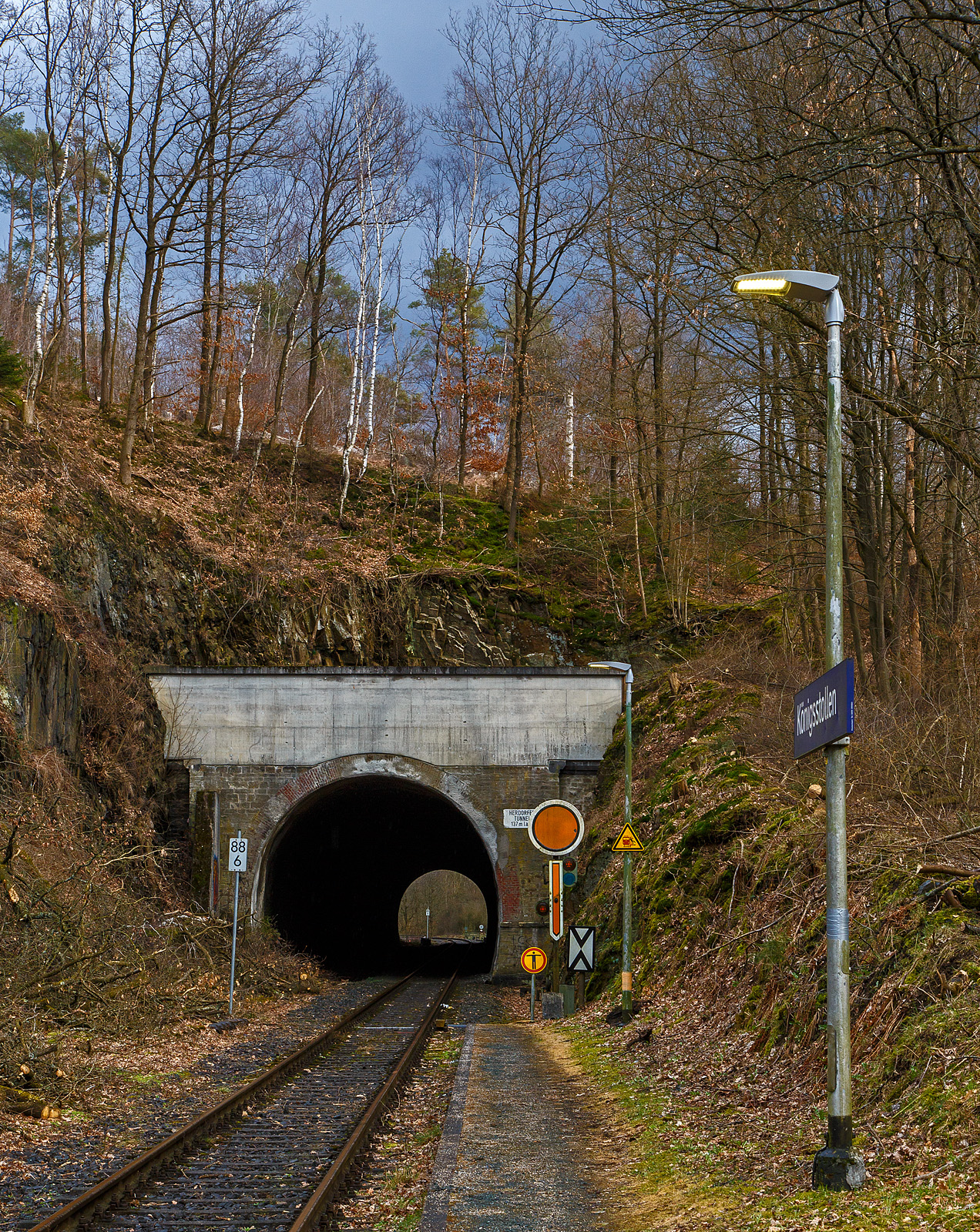 Der 137 m lange Herdorfer Tunnel hier am 18.03.2023 vom Hp Königsstollen gesehen. 

Davor das Vorsignal (fürs Einfahrtsignal Bf Herdorf) Vr 0 - Halt erwarten! Die gelbe Scheibe mit weißem Rand und gelbe Pfeil zeigt nach unten. Nachts leuchten auch die beiden gelben Lampen. Vor dem Signal die Ne 2 – Vorsignaltafel. Die Vorsignaltafel zeigt dem Lokführer, daß das Signal, vor dem sie steht, Vorsignalfunktion hat, d.h., den Begriff eines nachfolgenden Hauptsignales vorankündigt. 
