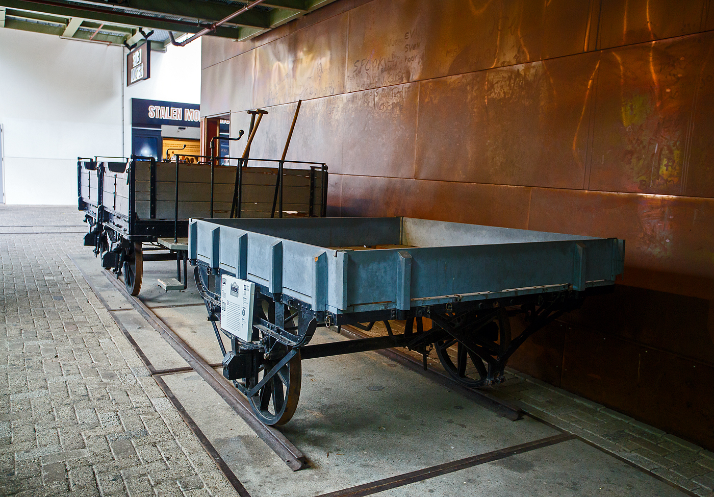 Der zweiachsige offene Pferdebahn-Güterwagen NTM G 1 und der einachsige offene Pferdebahn-Gepäckwagen NTM Dk 2 am 24 Juni 2025 im Het Spoorwegmuseum (bis 2005 NSM - Nederlands Spoorwegmuseum / Niederländischen Eisenbahnmuseum) in Utrecht im ehemaligen Bahnhof Maliebaan. Ein Eisenbahnmuseum das zum Verweilen einlädt. Übrigens Het vor Spoorwegmuseum heißt einfach „Das“, so heißt übersetzt das Museum einfach „Das Eisenbahnmuseum“. 

Die Wagen wurden beide von Beijnes, offiziell Koninklijke Fabriek van Rijtuigen en Spoorwagens J.J. Beijnes (Königliche Fabrik für Kutschen und Eisenbahnwagen J.J. Beijnes) in Haarlem gebaut und an die damalige NTM - Nederlandsche Tramweg Maatschappij (Niederländische Straßenbahngesellschaft) geliefert. Der NTM G 1 wurde 1880 und der NTM Dk 2 wurde 1881 gebaut. 

TECHNISCHE DATEN:
Gebaute Anzahl (Serie): 1 (G1) / 3 (Dk 2, Serie 1-3)
Spurweite: 1.435 mm (Normalspur)
Achsen: 2 (G1) / 1 (Dk 2)
Leergewicht: 600 kg (G1) / 300 kg (Dk 2)
Höchstgeschwindigkeit: 20 km/h

Die NTM:
Die NTM - Nederlandsche Tramweg Maatschappij (Niederländische Straßenbahngesellschaft) war ein Straßenbahn- und Busunternehmen, das zwischen 1880 und 1956 für den Bau, den Betrieb und die Instandhaltung von Straßenbahnlinien in Friesland verantwortlich war. Trotz ihres Namens war diese Straßenbahngesellschaft ausschließlich in Friesland und den angrenzenden Teilen tätig. Die letzte Tram fuhr 1956, als regionales Busunternehmen blieb die NTM bis 1971 aktiv.

Geschichte:
Die NTM wurde am 24. April 1880 in Utrecht gegründet, war aber später in Heerenveen ansässig. Ursprünglich plante das Unternehmen den Bau von Straßenbahnlinien in den Niederlanden, konzentrierte sich jedoch letztendlich auf Friesland und erweiterte das Angebot um einige benachbarte Provinzen.

Die letzten verbliebenen Straßenbahnlinien der ehemaligen NTM wurden 1956 an die NS (Niederländische Eisenbahnen) übertragen, die 1985 die letzte Straßenbahnlinie zwischen Drachten und Groningen, die damals nur noch dem Güterverkehr diente, stilllegte.

Pferdebahnen:
Die ersten von der NTM gebauten Linien wurden mit Pferdebahnen betrieben. Die erste Pferdebahnlinie führte von Dokkum nach Veenwouden und wurde am 6. Oktober 1880 eröffnet. Diese und alle nachfolgenden Linien waren in Normalspur (Spurweite 1.435 mm) ausgeführt. Ein Jahr später, am 19. September 1881, wurde die Strecke Veenwouden–Bergumerdam eröffnet, die 1896 bis Suameer verlängert wurde. 1898 wurde die Pferdebahnlinie Harkezijl–Makkum eröffnet, während die Pferdebahnlinie Berlikum–Sint Annaparochie schließlich 1900 eröffnet wurde. Diese letzte Linie wurde 1917 stillgelegt, während die Verbindung Harkezijl–Makkum bis zum 16. Juli 1930 in Betrieb blieb. Dies war die letzte Pferdebahnlinie in den Niederlanden.

Ab 1913 (Suameer–Veenwouden) und 1926 (Veenwouden–Dokkum) wurde die Pferdebahn durch die Dampfbahn ersetzt. Die Pferdebahnlinien waren leichter als andere Linien, daher mussten die Schienen durch schwerere ersetzt werden, bevor die schwereren Dampflokomotiven fahren konnten.

Dampfstraßenbahnen:
65 Jahre lang bildete die Dampfstraßenbahn den größten Teil der NTM, die in ihrer Blütezeit einen großen Teil der Provinz Friesland abdeckte. Die NTM war damit eine der größten Dampfstraßenbahngesellschaften der Niederlande.

Die erste Dampfstraßenbahn fuhr am 11. Januar 1882 auf der Straßenbahnlinie Sneek – Bolsward. Im selben Jahr kamen die Strecken Bolsward–Harlingen, Heerenveen–Joure und Heerenveen–Gorredijk hinzu, die 1884 bis nach Drachten verlängert wurden. 1886 wurde die fehlende Verbindung zwischen Joure und Sneek fertiggestellt. Zu diesem Zeitpunkt war die durchgehende Straßenbahnlinie Harlingen–Bolsward–Sneek–Joure–Heerenveen–Gorredijk–Drachten fertiggestellt. Zehn Jahre später, 1896, wurde die Straßenbahnlinie Veenwouden–Suameer nach Drachten verlängert. Diese Straßenbahnlinien hatten alle Normalspur, so dass Güterwagen von der Dampfstraßenbahn auf das nationale Eisenbahnnetz übertragen werden konnten.

Die Straßenbahnlinien dienten sowohl dem Personen- als auch dem Güterverkehr, wobei der Güterverkehr auf einigen Strecken den Personenverkehr um viele Jahre überdauerte. Das gesamte Netz bestand nur etwa 17 Jahre, da 1933 die letzte eröffnete Linie von Hijkersmilde entlang des Drentsche Hoofdvaart-Kanals nach Meppel als erste stillgelegt wurde. 

Am 13. August 1939 wurden mehrere Verbindungen eingestellt. Die verbleibenden Straßenbahnlinien blieben bis nach dem Zweiten Weltkrieg in Betrieb, doch 1947 wurde der größte Teil des Netzes stillgelegt. 

Lediglich die letzte Linie Drachten – Groningen blieb noch längere Zeit in Betrieb. Das in den 1950er Jahren in Drachten gegründete Philips-Werk war sein Hauptkunde. Bis in die 1980er Jahre wurde auf der Philips-Linie durch die NS regelmäßiger Güterverkehr mit Diesellokomotiven der Baureihe NS 2400 betrieben. 1973, 1975 und 1976 kehrte die Dampfstraßenbahn zurück, als die Dampfstraßenbahn Hoorn-Medemblik (SHM) auf dieser alten Straßenbahnlinie verkehrte. Die letzte Fahrt auf der gesamten Strecke erfolgte mit Wadlopern am 30. April 1985, eine weitere Fahrt nach Roden am 7. Juni 1985. 1988 wurde die Strecke stillgelegt. Dies markierte das Ende der Geschichte der friesischen Straßenbahnlinien.
