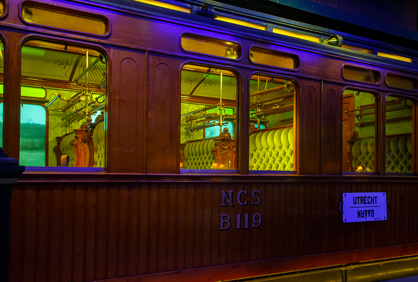 Detailbild des zweite Klasse Reisezugwagen NCS B 119, ein luxuriöser Teakholzwagen der ehemaligen NCS - Nederlandsche Centraal Spoorwegmaatschappij (Niederländische Zentralbahngesellschaft) am 24. Juni 2025 im Het Spoorwegmuseum (bis 2005 NSM - Nederlands Spoorwegmuseum / Niederländischen Eisenbahnmuseum) in Utrecht im ehemaligen Bahnhof Maliebaan. Nicht nur die Compagnie Internationale des Wagons-Lits (CIWL) für ihre Luxuszüg, wie beispielsweise dem Orient-Express, hatte solche Wagen.

Dieser äußerst luxuriösen Abteilwagen vom Typ BL8 (Serie B 111 – 127) wurde 1904 in der eigenen Werkstatt der NCS in Utrecht (Werkplaats NCS) gebaut. 

Bedingt durch den Ersten Weltkrieg wurde 1917 eine Zweckgemeinschaft der unabhängigen Eisenbahngesellschaften HSM beziehungsweise HIJSM (Hollandsche IJzeren Spoorweg-Maatschappij) und SS (Maatschappij tot Exploitatie van Staatsspoorwegen) gegründet. Da die NCS unter der Kontrolle der SS stand, wurde sie von diesem Moment an auch Teil der neuen Partnerschaft. Der Betrieb wurde am 1. Juni 1919 an die SS übertragen. Die beiden Gesellschaften errichteten einen gemeinsamen Bahnbetrieb, blieben aber rechtlich unabhängig. Dies änderte sich 1938, als HIJSM und SS vollständig fusionierten und in der AG NS (Nederlandse Spoorwegen, früher Nederlandsche Spoorwegen geschrieben) aufgingen. Das Rollmaterial wurde bereits 1921 zusammengelegt. Somit wurde ab 1924 der Wagen als NS B 1814 geführt, 1952 wurde er außer Dienst gestellt und für das Eisenbahnmuseum erhalten. Er kam 1977 vollständig restauriert als NCS B 119 dort an. Seitdem ist ein Paradewagen des Museums.

Anfang des 20. Jahrhundert war es den lokalen Eisenbahnen in den Niederladen gesetzlich verboten Waggons der 1. Klasse einzusetzen. Um doch wohlhabende Passagiere von Zeist und Baarn (einschließlich des Direktors der NCS) in passender Art zu befördern umging die NCS mit diesen als 2. Klasse Wagen klassifizierten äußerst luxuriösen Standard. Neben den teakholzgetäfelten Außenwänden ist auch der Innenraum opulent und gepflegt. Zu beiden Seiten des Mittelgangs befinden sich gepolsterte Doppelbänke mit Wendekissen. Im Winter war die plüschige Seite oben, im Sommer die kühlere, lederbezogene Seite. Lampen, Gepäckablagen, Aschenbecher, alles ist wunderschön verarbeitet und aus Kupfer gefertigt. Die Waggons wurden unter anderem auf den (Lokal-)Bahnen Utrecht–Den Dolder–Baarn und Utrecht–Bilthoven–Zeist eingesetzt. Insgesamt gab es siebzehn dieser Wagen.

