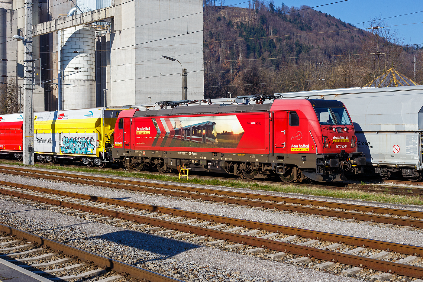 Die an die Stern & Hafferl Verkehrsgesellschaft m.b.H. (StH) vermiete 187 333-0 „Barbara“ (91 80 6187 333-0 D-Rpool, ex 91 81 1187 333-0 A-STH) der Railpool GmbH (München) kommt am 03 April 2025 im Dieselbetrieb, mit einem Kalkstein beladenen RockTainer SAND-Zug, vom Kalksteinbruch in Ebensee kommend beim Bahnhof Gmunden bzw. beim Zementwerk Hatschek der Rohrdorfer Zement GmbH in Gmunden an.

Die Bombardier TRAXX F140 AC3 LM wurde 2018 von der Bombardier Transportation GmbH in Kassel unter der Fabriknummer 35513 gebaut und an die Stern & Hafferl Verkehrsgesellschaft m.b.H. (Gmunden) geliefert. Die Lok wurde 2023 an Railpool verkauft und STH hat sie seitdem nur noch gemietet. Sie hat die Zulassungen für Deutschland (D) und Österreich (A), für H/RO sind durchgestrichen. Fragwürdig ist die angeschriebene UIC-Nummer …D-Rpool, da es nicht mit den Nummern übereinstimmt.  

Der Zug bestand aus 2 x 4-achsigen Flachwagen in Sonderbauart (Doppelwagen) bzw. 8-achsig Containertragwagen, der Gattung Sggrrs 80 ft, der österreichischen INNOFREIGHT bzw, Rail Cargo Austria. Die achtachsigen Wageneinheiten (80ft Sggrrs IW) sind für den Transport von Ladegut mit Ladegestellen geeignet, hier in der Variante J - RockTainer SAND mit einer ISO-Länge von 40‘. Mit einer maximalen Zuladung von 134,4 Tonnen pro Doppelwagen können große Mengen problemlos transportiert werden. Das ist mit herkömmlichen Containern oder auf LKWs unvorstellbar. Die Entladung erfolgt in einen Tiefbunker.

Die Rohrdorfer Unternehmensgruppe nutzt für die Lieferung von Kalkstein vom Steinbruch Ebensee in das Zementwerk Gmunden die neuen RockTainer SAND-Tragwagen der österreichischen ÖBB Rail Cargo Group (RCG), dem führenden Bahnlogistiker in Europa. Die Spezialtransportwaggons wurden von der RCG in Zusammenarbeit mit der Rohrdorfer Unternehmensgruppe und Innofreight, ein österreichisches Gütertransport- und Logistikunternehmen, entwickelt, um mehr Ladung mit weniger Fahrten transportieren zu können. Seit Mai 2023 werden die 20 neuen, sogenannten InnoWaggons mit 40 RockTainer SAND-Containern auf der ca. 15 km langen Strecke Ebensee–Gmunden (Teilstück der „Salzkammergutbahn“ (ÖBB 170) Bahnstrecke Stainach-Irdning über Attnang-Puchheim nach Schärding). eingesetzt. Insgesamt werden in diesem Verkehr je Transportperiode rund 450.000 Tonnen Kalkstein befördert. Das sind 36.000 Lkw-Fahrten, die jedes Jahr dank der umweltfreundlichen Schiene und des innovativen Equipments eingespart werden können.
