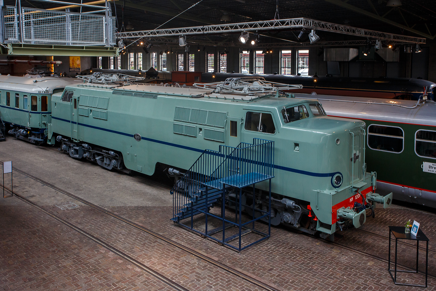 Die Elektrolokomotive NS 1201 der NS-Baureihe 1200 am 24 Juni 2025 im Het Spoorwegmuseum (bis 2005 NSM - Nederlands Spoorwegmuseum / Niederländischen Eisenbahnmuseum) in Utrecht im ehemaligen Bahnhof Maliebaan. Ein Eisenbahnmuseum das zum Verweilen einlädt.

Die Lok wurde 1951 von Werkspoor in Utrecht unter der Fabriknummer 928 gebaut (Baldwin-Lizenz) und an die NS - Nederlandse Spoorwegen (Niederländischen Staatsbahnen, heute Niederländische Eisenbahnen AG) geliefert. Wobei die Entwicklung und Konstruktion, sowie auch einige Teile, von Baldwin Locomotive Works in Philadelphia (USA) stammt. Auch die Entwicklung der Elektrotechnik stammt aus den Vereinigten Staaten und zwar von Westinghouse Electric Corporation (Monroeville), diese wurde von Heemaf in Hengelo (Westinghouse-Lizenz) geliefert.

Die Baureihe 1200 der Niederländischen Eisenbahnen (NS) ist eine sechsachsige Elektrolokomotivbaureihe aus den Jahren 1951 bis 1953. Die Lokomotiven waren bis 1998 bei der NS im Einsatz.

Die Nase war der erste Teil dieser unglaublich starken und robusten Lokomotive, der ins Auge fiel. Sie sollte dem Lokführer bei Kollisionen einen besseren Schutz bieten. Diese Lokomotiven wurden in den Niederlanden nach einem amerikanischen Design und mit einigen amerikanischen Teilen gebaut, die teilweise durch amerikanische Dollar über den Marshall-Plan finanziert wurden. Die NS nutzte diese Lokomotiven, um sowohl Personenzüge als auch Güterzüge zu ziehen. Ihre wichtigste Aufgabe war es Schnellzüge zu ziehen. Als sich die NS 1995 trennte, wurden alle 1200er Jahre an NS Cargo übertragen und begannen im Güterverkehr zu arbeiten. 

Nach Ende des Zweiten Weltkrieges gab es großen Fahrzeugmangel bei der NS. Da man keine Erfahrungen mit dem Bau elektrischer Lokomotiven hatte, kaufte man Lokomotiven bei verschiedenen Lieferanten. Die bei Baldwin konstruierten Lokomotiven wurden bei Werkspoor in Utrecht gefertigt. Die bei Westinghouse entwickelte Elektrotechnik wurde von Heemaf in Hengelo geliefert. Von den ursprünglich 75 georderten Lokomotiven wurden nur 25 ausgeliefert. Der amerikanische Einfluss ist an den hochliegenden Führerständen und den großen, dem Schutz des Lokführers dienenden Vorbauten erkennbar. Am 30. November 1950 wurden 25 Loks bestellt, am 27. September 1951 konnte die erste Probefahrt stattfinden. Am 9. Februar 1952 wurde die 1201 offiziell in Dienst gestellt, der Preis pro Lok betrug 765.000 NFL (ca. 347.000 Euro).

Die Loks hatten sechs Motoren des Typs TM94. Die ersten 14 Lokomotiven waren türkis lackiert, danach setzte sich wegen des sichtbaren Abriebs der Stromabnehmer eine rotbraune Farbgebung durch. Ab 1954 erhielten alle Loks eine blaue Farbgebung, 1971 erhielt die erste Lok die neuen NS-Hausfarben grau/gelb, 1983 war auch die letzte Lok entsprechend umlackiert. Am 30. März 1998 endete der planmäßige Einsatz bei der NS.

Fünf weitere Lokomotiven (1214, 1215, 1218, 1221, 1225) wurden 1999 an die ACTS Nederland BV verkauft. Diese vermietete 2010 drei Lokomotiven (1214, 1215, 1225) an die Eisenbahngesellschaft Euro-Express Treincharter (EETC). Von der wurden sie bis April 2015 unter anderem vor den Nachtzügen Niederlande–Österreich zwischen Amsterdam und Venlo eingesetzt. Fünf Maschinen (1201, 1202, 1211, 1218, 1221) sind als Museumslokomotiven erhalten. NS 1211 ist Botschafterlok im Bahnpark Augsburg. Leihgeber ist das Eisenbahnmuseum Utrecht.

TECHNISCHE DATEN:
Hersteller mech.: Werkspoor (in Lizenz von Baldwin)
Hersteller Elektrik: Heemaf (in Lizenz von Westinghouse)
Baujahre: 1950 bis 1953
Ausmusterung bei NS: bis 1998 
Gebaute Anzahl: 25
Nummerierung: NS 1201 bis 1225
Spurweite: 1.435 mm (Normalspur)
Achsfolge: Co’Co’ 
Länge über Puffer: 18.086 mm
Höhe: 4.064 mm
Treibraddurchmesser: 1.100 mm (neu)
Dienstgewicht: 108 t
Antriebsmotoren: 6 Stück Heemaf vom Typ TM 94
Getriebeübersetzung: 20 : 71
Höchstgeschwindigkeit: 130 km/h (Planmäßig) / 150 km/h (technisch möglich)
Stundenleistung: 6 x 392 kW = 2.350 kW
Dauerleistung: 6 x 367 kW = 2.200 kW
Anfahrzugkraft: 195 kN
Dauerzugkraft: 164 kN bei 80 km/h / 64,7 kN bei 123 km/h
Stromsystem: 1,5 kV DC (=)
Kleister befahrbarer Gleisbogen: R 100 m