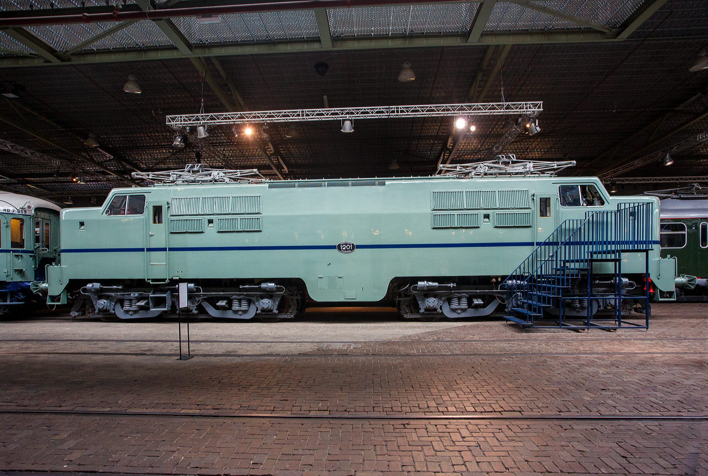 Die Elektrolokomotive NS 1201 der NS-Baureihe 1200 am 24 Juni 2025 im Het Spoorwegmuseum (bis 2005 NSM - Nederlands Spoorwegmuseum / Niederländischen Eisenbahnmuseum) in Utrecht im ehemaligen Bahnhof Maliebaan. Ein Eisenbahnmuseum das zum Verweilen einlädt.

Die Lok wurde 1951 von Werkspoor in Utrecht unter der Fabriknummer 928 gebaut (Baldwin-Lizenz) und an die NS - Nederlandse Spoorwegen (Niederländischen Staatsbahnen, heute Niederländische Eisenbahnen AG) geliefert. Wobei die Entwicklung und Konstruktion, sowie auch einige Teile, von Baldwin Locomotive Works in Philadelphia (USA) stammt. Auch die Entwicklung der Elektrotechnik stammt aus den Vereinigten Staaten und zwar von Westinghouse Electric Corporation (Monroeville), diese wurde von Heemaf in Hengelo (Westinghouse-Lizenz) geliefert.

Die Baureihe 1200 der Niederländischen Eisenbahnen (NS) ist eine sechsachsige Elektrolokomotivbaureihe aus den Jahren 1951 bis 1953. Die Lokomotiven waren bis 1998 bei der NS im Einsatz.

Die Nase war der erste Teil dieser unglaublich starken und robusten Lokomotive, der ins Auge fiel. Sie sollte dem Lokführer bei Kollisionen einen besseren Schutz bieten. Diese Lokomotiven wurden in den Niederlanden nach einem amerikanischen Design und mit einigen amerikanischen Teilen gebaut, die teilweise durch amerikanische Dollar über den Marshall-Plan finanziert wurden. Die NS nutzte diese Lokomotiven, um sowohl Personenzüge als auch Güterzüge zu ziehen. Ihre wichtigste Aufgabe war es Schnellzüge zu ziehen. Als sich die NS 1995 trennte, wurden alle 1200er Jahre an NS Cargo übertragen und begannen im Güterverkehr zu arbeiten. 

Nach Ende des Zweiten Weltkrieges gab es großen Fahrzeugmangel bei der NS. Da man keine Erfahrungen mit dem Bau elektrischer Lokomotiven hatte, kaufte man Lokomotiven bei verschiedenen Lieferanten. Die bei Baldwin konstruierten Lokomotiven wurden bei Werkspoor in Utrecht gefertigt. Die bei Westinghouse entwickelte Elektrotechnik wurde von Heemaf in Hengelo geliefert. Von den ursprünglich 75 georderten Lokomotiven wurden nur 25 ausgeliefert. Der amerikanische Einfluss ist an den hochliegenden Führerständen und den großen, dem Schutz des Lokführers dienenden Vorbauten erkennbar. Am 30. November 1950 wurden 25 Loks bestellt, am 27. September 1951 konnte die erste Probefahrt stattfinden. Am 9. Februar 1952 wurde die 1201 offiziell in Dienst gestellt, der Preis pro Lok betrug 765.000 NFL (ca. 347.000 Euro).

Die Loks hatten sechs Motoren des Typs TM94. Die ersten 14 Lokomotiven waren türkis lackiert, danach setzte sich wegen des sichtbaren Abriebs der Stromabnehmer eine rotbraune Farbgebung durch. Ab 1954 erhielten alle Loks eine blaue Farbgebung, 1971 erhielt die erste Lok die neuen NS-Hausfarben grau/gelb, 1983 war auch die letzte Lok entsprechend umlackiert. Am 30. März 1998 endete der planmäßige Einsatz bei der NS.

Fünf weitere Lokomotiven (1214, 1215, 1218, 1221, 1225) wurden 1999 an die ACTS Nederland BV verkauft. Diese vermietete 2010 drei Lokomotiven (1214, 1215, 1225) an die Eisenbahngesellschaft Euro-Express Treincharter (EETC). Von der wurden sie bis April 2015 unter anderem vor den Nachtzügen Niederlande–Österreich zwischen Amsterdam und Venlo eingesetzt. Fünf Maschinen (1201, 1202, 1211, 1218, 1221) sind als Museumslokomotiven erhalten. NS 1211 ist Botschafterlok im Bahnpark Augsburg. Leihgeber ist das Eisenbahnmuseum Utrecht.

TECHNISCHE DATEN:
Hersteller mech.: Werkspoor (in Lizenz von Baldwin)
Hersteller Elektrik: Heemaf (in Lizenz von Westinghouse)
Baujahre: 1950 bis 1953
Ausmusterung bei NS: bis 1998 
Gebaute Anzahl: 25
Nummerierung: NS 1201 bis 1225
Spurweite: 1.435 mm (Normalspur)
Achsfolge: Co’Co’ 
Länge über Puffer: 18.086 mm
Höhe: 4.064 mm
Treibraddurchmesser: 1.100 mm (neu)
Dienstgewicht: 108 t
Antriebsmotoren: 6 Stück Heemaf vom Typ TM 94
Getriebeübersetzung: 20 : 71
Höchstgeschwindigkeit: 130 km/h (Planmäßig) / 150 km/h (technisch möglich)
Stundenleistung: 6 x 392 kW = 2.350 kW
Dauerleistung: 6 x 367 kW = 2.200 kW
Anfahrzugkraft: 195 kN
Dauerzugkraft: 164 kN bei 80 km/h / 64,7 kN bei 123 km/h
Stromsystem: 1,5 kV DC (=)
Kleister befahrbarer Gleisbogen: R 100 m