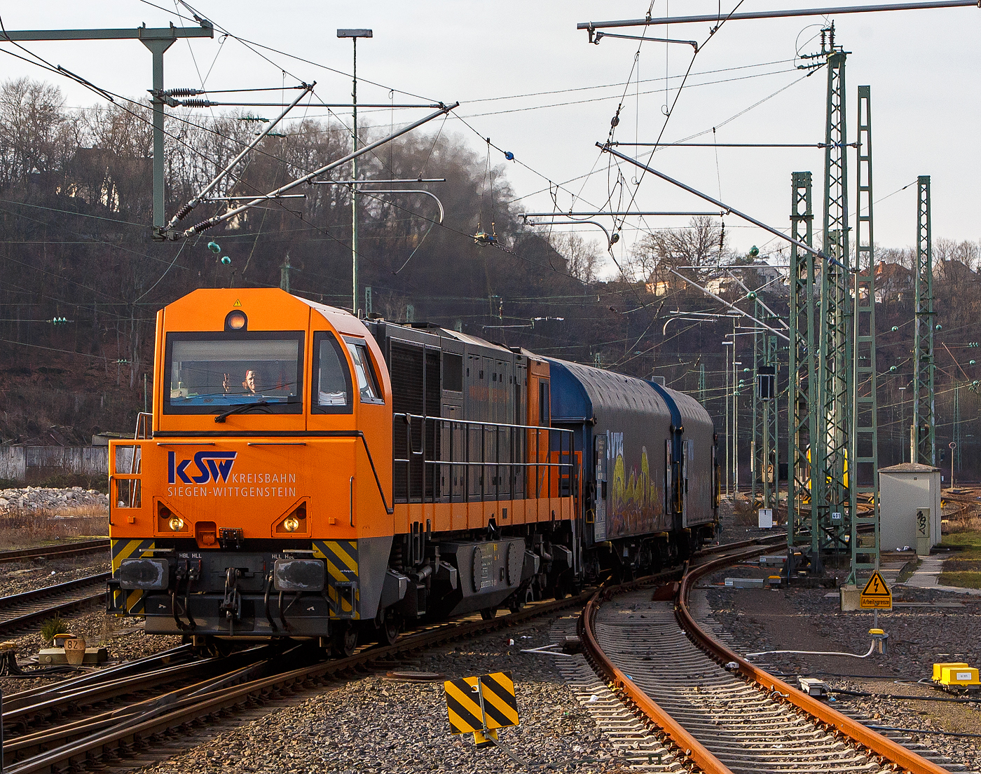 Die KSW 43 (92 80 1273 018-2 D-KSW), die asymmetrische Vossloh G 2000 BB der KSW (Kreisbahn Siegen-Wittgenstein) fährt am 09.02.2023, mit ihrem Coilzug (VTG Coilwagen der Gattung Shimmns-ttu), von Betzdorf (Sieg) über Siegen, Dillenburg und Haiger nach Herdorf, was einem Umweg von über 62 km bedeutet. (Aufnahme vom Bahnsteig 106)

Eigentlich würde der Zug hier auf die KBS 462 – Hellertalbahn abbiegen und er wäre in 7,2 km am Ziel. Aber die Hellertalbahn bleibt, nach dem Felssturz am 23.12.2022 in Herdorf bis voraussichtlich zum kleinen Fahrplanwechsel am 09. Juni 2023 gesperrt. Ein weiterer Nachteil ist das die Strecke Haiger – Herdorf nur die Streckenklasse CE hat und zudem sehr Steigungsreich ist. Die 7,2 km lange Strecke Betzdorf – Herdorf hat die Streckenklasse D4, so können die Coilwagen auch nur abgelastet zugestellt werden.

Nochmal einen lieben Gruß an den netten Lokführer zurück.

Zudem habe ich es hier nicht verstanden, warum der KSW-Zug erst auf den Regionalverkehr warten musste bevor es weiter ging. Denn der Zug könnte doch ohne Halt durchfahren.
