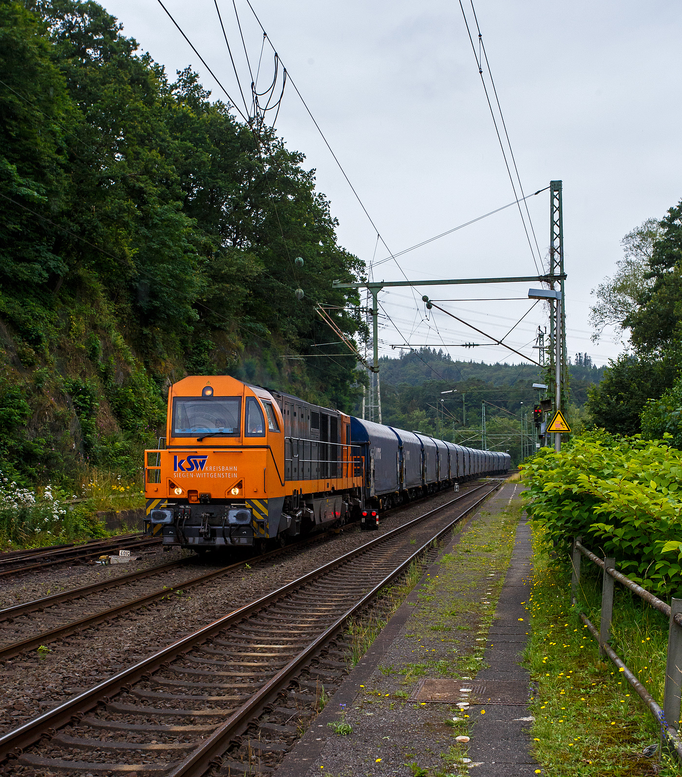 Die KSW 43 (92 80 1273 018-2 D-KSW), die asymmetrische Vossloh G 2000 BB der KSW (Kreisbahn Siegen-Wittgenstein) fährt am 03 Juli 2024, mit einem beladen Coilzug (16 Stück VTG Coilwagen der Gattung Shimmns-ttu, VTG Bez. F46.520D) vom Hafen Duisburg kommend, durch den Bahnhof Scheuerfeld(Sieg) in Richtung Herdorf via Betzdorf. 

In Betzdorf (Sieg) geht es dann über KBS 462 „Hellertalbahn“ nach Herdorf zum Rbf der KSW (Betriebsstätte FGE -Freien Grunder Eisenbahn). Für die Weiterfahrt auf den Pfannenberg (in Neunkirchen-Salchendorf) über nimmt dort dann die KSW 47 (92 80 1271 027-5 D-KSW), ex D 2 der HFM, eine Vossloh G 1000 BB, den Zug.
