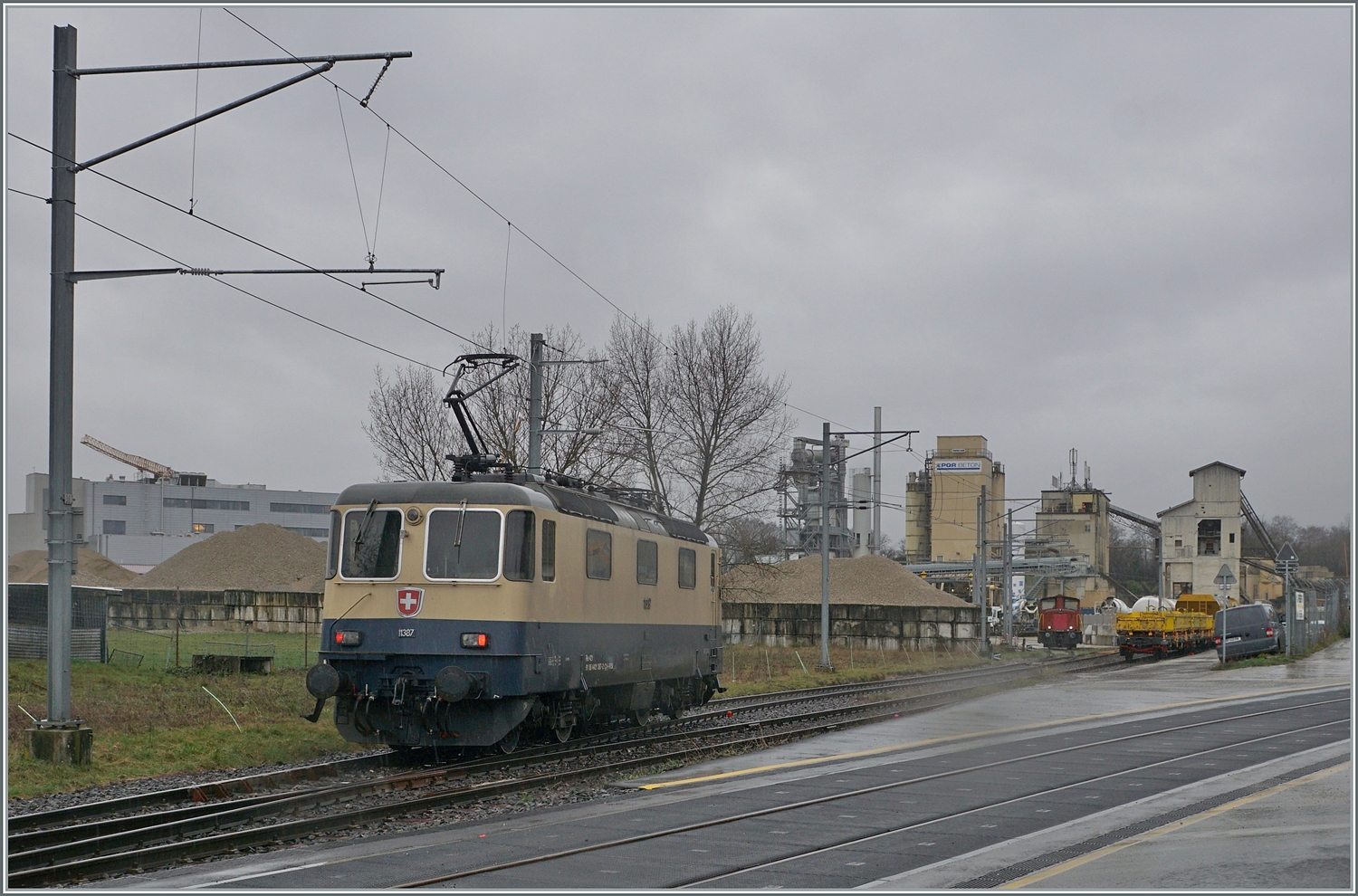 Die von Morges gekommene IRSI/IGE  Rheingold  Re 4/4 II 11387 (Re 421 387-2) hat in Gland die Strecke nach Genève verlassen und fährt nun über ein Industriegleis in Richtung Kieswerk, um dort ihren Zug nach Apples abzuholen. 
Die angemietete IRSI/IGE Re 4/4 II 11387 (Re 421 387-2) ersetzt die zur Zeit in der Revision in Bellinzona weilende BAM MBC MBC Re 420 506. 

22. Feb. 2024