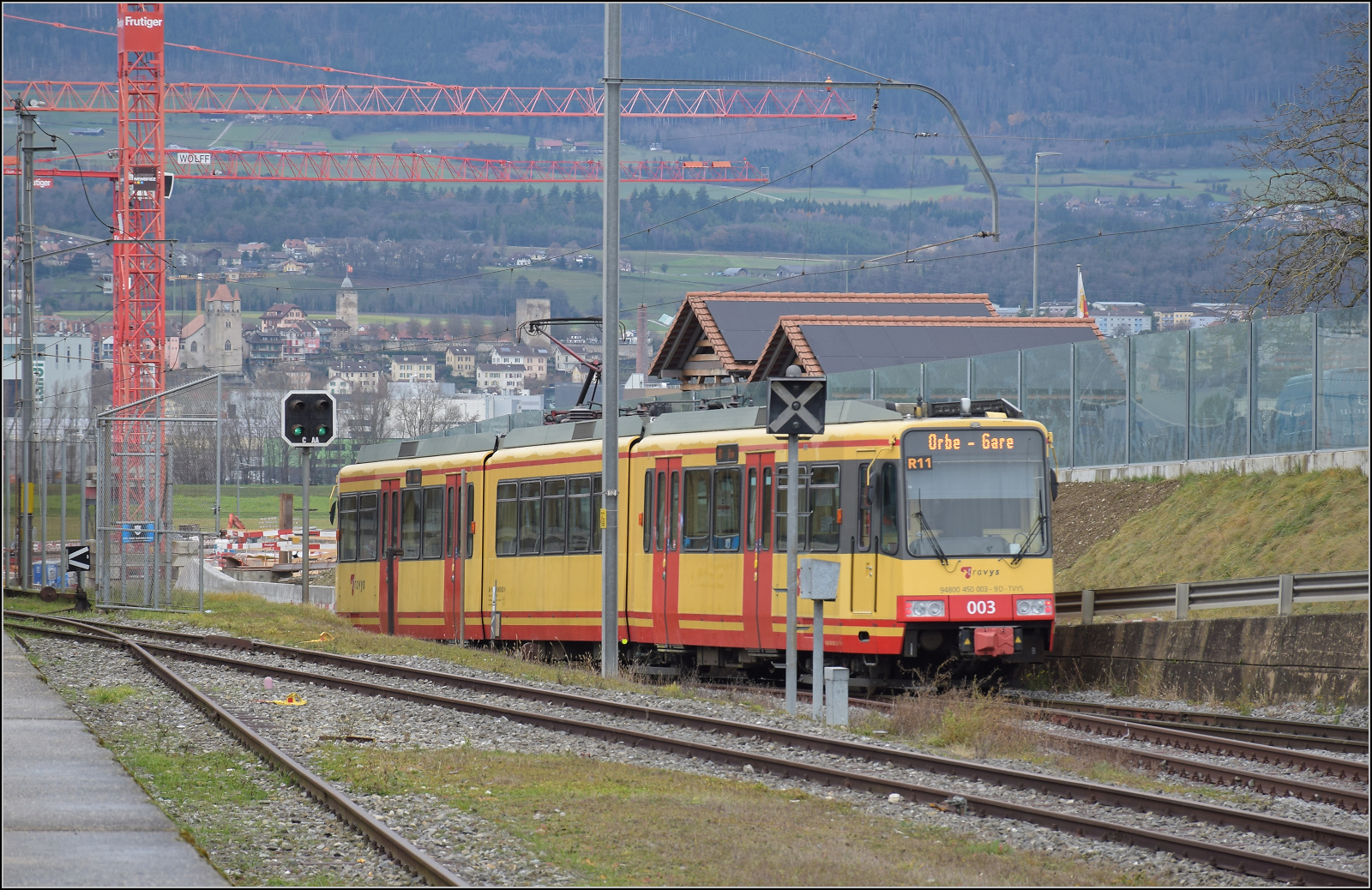 Ende einer Ära - der Gleichstrom und die  Lokalbahn  gehen. 

Die Karlsruher Zweisystem-Strassenbahn  Be 4/8 003  alias 94 800 450 003-9 D-TVYS fährt nach Orbe aus Chavornay aus. Eigentlich ein misslungenes Bild zum Aussortieren. Und doch zeigt es die Geschichte der verschwindenden und schon leicht verschwimmenden Bahn im Gleichstrommodus auf dem bald nicht mehr benutzten Streckenteil. Die Kräne für den Brückenbau zur Vermeidung eines Bahnübergangs sind bereits unübersehbar vor der historischen Altstadkulisse von Orbe aufgebaut. Dezember 2025.