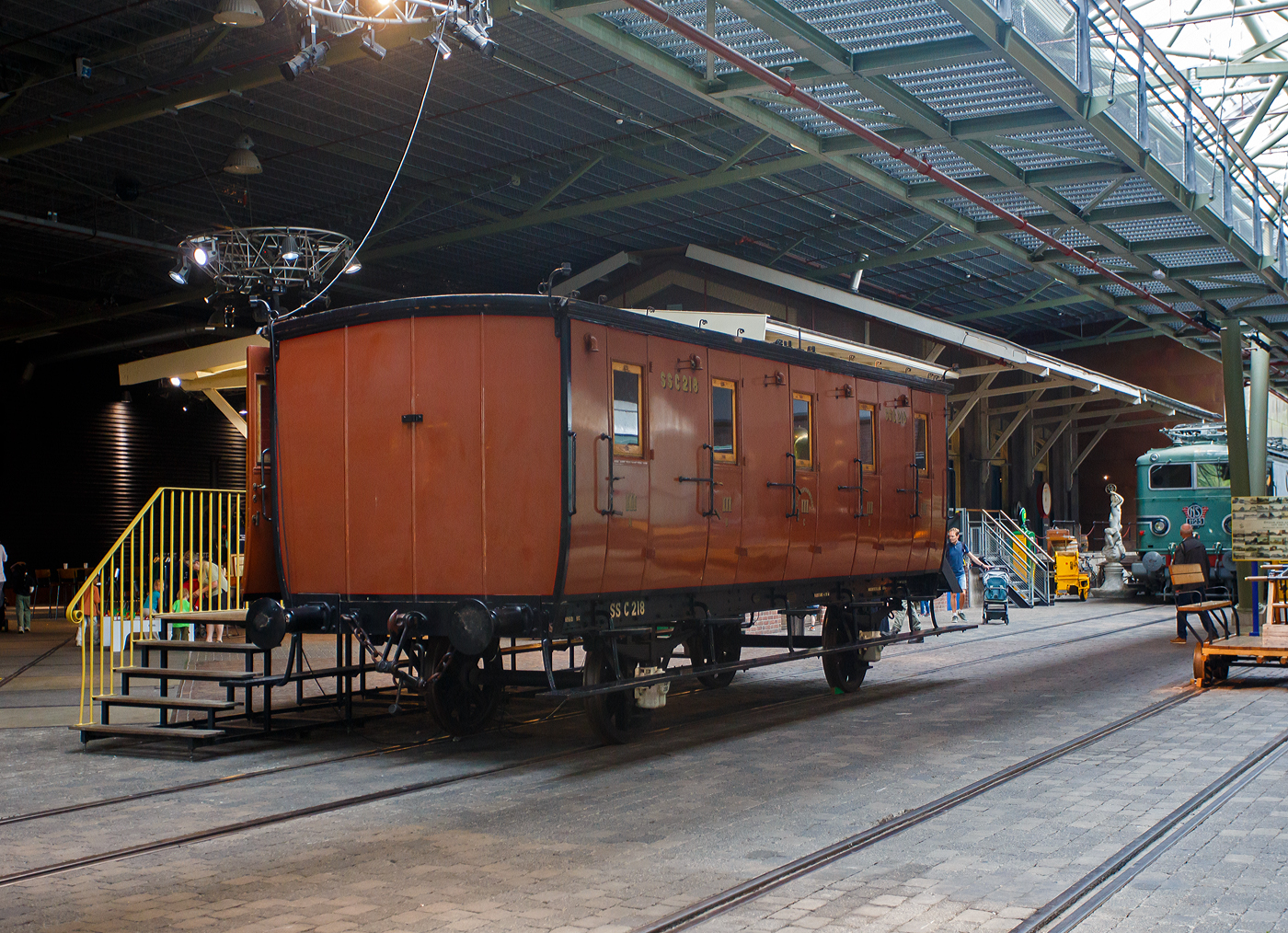 SS C 218
Der zweiachsige dritte Klasse Reisezugwagen SS C 218 „Koekblik“ (Keksdose), Baujahr 1874, der älteste erhaltene Personenwagen der Niederlande, am 24 Juni 2025 im Het Spoorwegmuseum (bis 2005 NSM - Nederlands Spoorwegmuseum / Niederländischen Eisenbahnmuseum) in Utrecht im ehemaligen Bahnhof Maliebaan. Im Museum wird er wegen seiner Form und der Metallverkleidung an der Außenseite „Koekblik“ (Keksdose) genannt. 

Der Wagen wurde 1874 von Beijnes, offiziell Koninklijke Fabriek van Rijtuigen en Spoorwagens J.J. Beijnes (Königliche Fabrik für Kutschen und Eisenbahnwagen J.J. Beijnes) in Haarlem gebaut und an die damalige SS - Maatschappij tot Exploitatie van Staatsspoorwegen (Gesellschaft zum Betrieb von Staatseisenbahnen) geliefert. Er gehörte zu einer Serie von 339 Exemplaren des Typs C5 – 3.Klasse Reisezugwagen mit 5 Abteilen (Serie C 1 - 339) die gebaut wurden. Dieser Wagen ist das einzige noch existierende Exemplar dieser Serie und damit der älteste erhaltene Reisezugwagen der Niederlande. 

Diese Waggons ähnelten noch immer stark an seinen Vorgänger, der Postkutsche. Die Wagen waren nicht besonders komfortabel, der Transport steht im Vordergrund. Er hat keine Heizung, und die Bänke sind aus Holz. Es gibt separate Abteile mit Türen, die nur von außen geöffnet werden können, und einer Außentrittstufe für den Schaffner. Jedes Abteil der 5 Abteile bietet Platz für 10 Personen. Es gibt auch ein separates Damenabteil für allein reisende Frauen. Die Rückenlehnen der Holzbänke reichten nicht alle bis zur Decke. Dadurch konnte der gesamte Waggon mit nur zwei Öllampen beleuchtet werden. 1892 wurde der Waggon mit Gasbeleuchtung ausgestattet, kurz darauf folgte der Einbau einer Heizung, diese wurde mit Dampf aus der Lokomotive betrieben.

Auf dem Dach des Waggons befinden sich wie ein Schweineschwanz geformte Haken sogenannte „Pigtails“, durch die wurde ein Seil, entlang der gesamten Zuglänge geführt, das mit einer Glocke am Tender der Lokomotive verbunden war. So konnte der Schaffner den Lokführer im Notfall warnen bzw. vereinbarte Signale geben. 

Die Wagen waren übrigens nicht alle exakt identisch, es gab Maßabweichungen. Die späteren Wagen hatten mehr Fenster pro Abteil. Jedes Abteil bot Platz für zehn Reisende auf Holzbänken. Es war ein Standardtyp der SS, aber dieselbe Konstruktion fand sich auch bei der NBDS und der NCS. Auch die AB-Wagen (1. und 2. Klasse) verwendeten dasselbe Fahrgestell, aber hier hatten die Reisenden mehr Platz, weil nur vier Abteile auf dem Fahrgestell angebracht waren. Außerdem saßen sie auf gepolsterten Bänken. Die NBDS hatte solche Wagen sogar für die 4. Klasse, die auf der NBDS-Strecke von Gennep nach Gogh in Deutschland im Einsatz waren. Diese einfachen Wagen hatten nur 15 Sitzplätze und 35 Stehplätze. Die 4. Klasse wurde in Deutschland 1928 eingestellt.

Die 1921 noch vorhandenen ehemaligen SS-Wagen erhielten von der NS die Nummern C 2126–2285 (C5). Ab 1917 bilden die Eisenbahngesellschaften HSM beziehungsweise HIJSM (Hollandsche IJzeren Spoorweg-Maatschappij) und SS (Maatschappij tot Exploitatie van Staatsspoorwegen) eine Zweckgemeinschaft, ab 1921 wurde das Rollmaterial, als NS - Nederlandsche Spoorwegen zusammengelegt. Die C 2126 bis 2285 Wagen wurden zwischen 1931 und 1933 größtenteils verschrottet.

Der Wagen SS C 218 wurde jedoch bereits 1922 in einen Dienstwagen des Straßen- und Bauamts mit der Nummer NS 177007 umgebaut. Diese Funktion behielt der Wagen bis 1952. Zunächst drohte ihm auch der Abriss, er wurde jedoch aus historischen Gründen erhalten. Und so blieb er der einzige erhaltene Wagen dieser Serie. Schließlich übernahm die Wagenwerkstatt in Amersfoort den alten Wagen und restaurierte ihn in seinen ursprünglichen Zustand. Nach der Fertigstellung im Jahr 1975 wurde er dem Eisenbahnmuseum gespendet. Im Museum wird sie aufgrund ihrer Form und der Metallverkleidung an der Außenseite „Koekblik“ / „Keksdose“ genannt. 

TECHNISCHE DATEN:
Baujahr: 1874
Hersteller: Beijnes, Haarlem
Gebaute Anzahl: 339 (Typ C5 - 3.Klasse Reisezugwagen mit 5 Abteilen).
Spurweite: 1.435 mm (Normalspur)
Achsen: 2
Länge über Puffer: 9.190 mm
Achsabstand: 4.700 mm
Eigengewicht: 8.960 kg
Höchstgeschwindigkeit: 75 km/h
Abteile: 5 á 10 Personen  
Sitzplätze: 50 (in der 3.Klasse)

Quellen: Het Spoorwegmuseum, nmld.nl und Anschriften
Stand/Überarbeitung: September 2025
